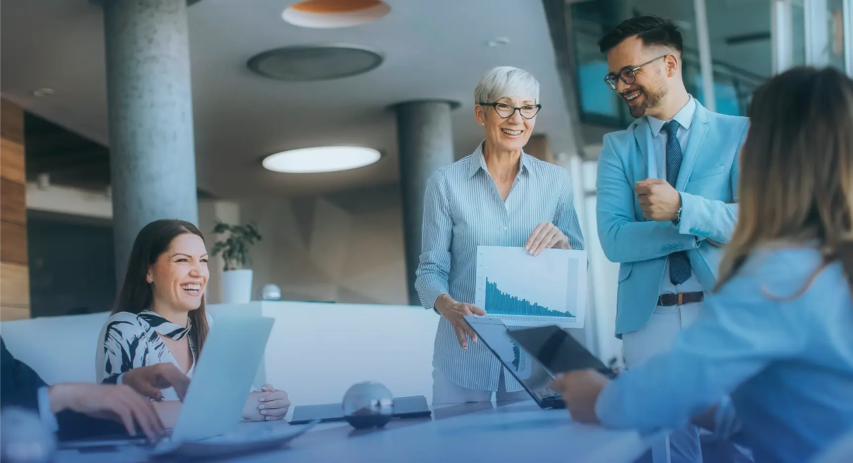 Grupo de profesionales en reunión de oficina, mujer mayor mostrando gráfico en papel mientras colegas sonríen y conversan.
