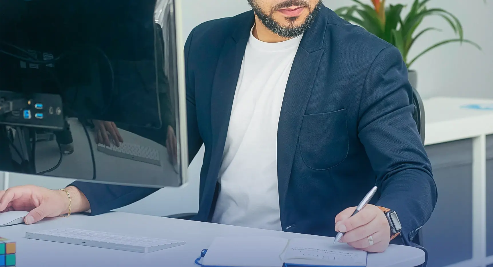 Hombre con saco azul trabajando en un escritorio mientras escribe en una libreta y usa la computadora.