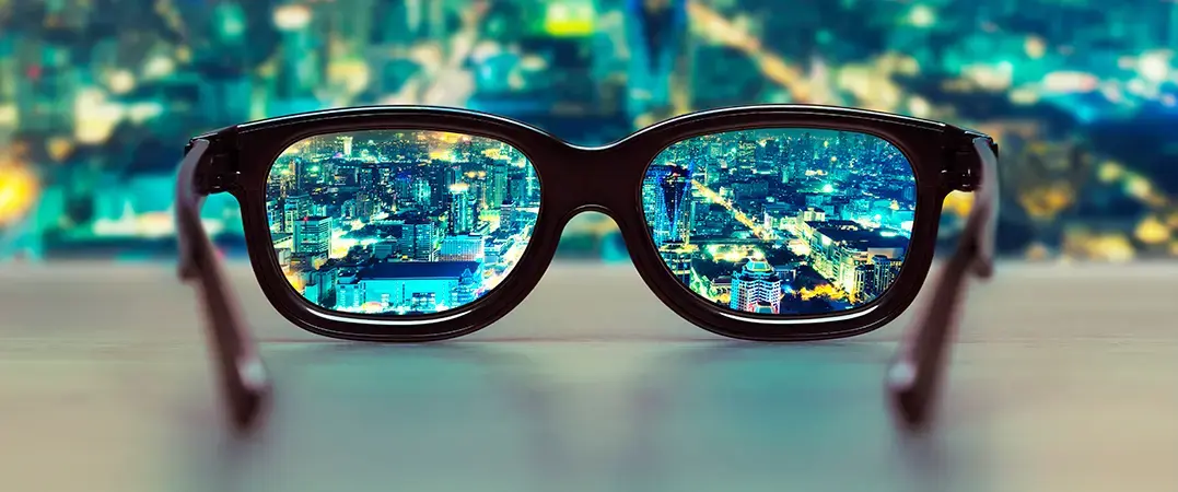 Vista de una ciudad nocturna reflejada con nitidez a través de unos lentes sobre una superficie, con el fondo desenfocado.