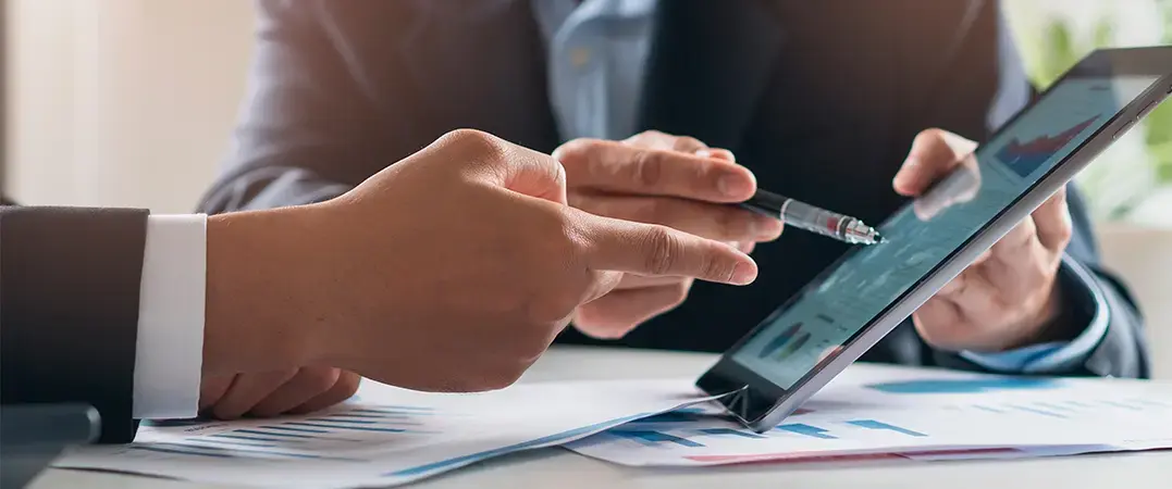 Dos personas en traje revisando gráficos financieros en una tablet sobre una mesa con documentos.